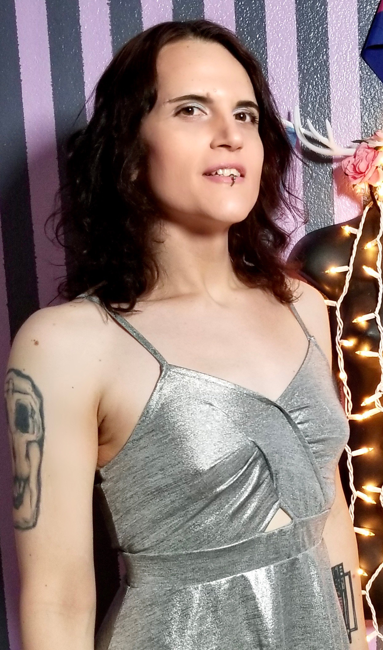 A headshot of Meika Grimm wearing a silver dress. She has long dark hair and is smiling in front of a decorative wall with lights